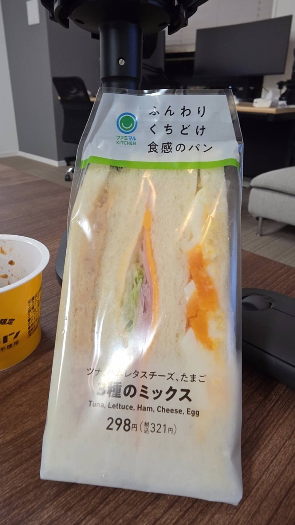 机の上に置かれた、ファミリーマートの「5種のミックス」サンドイッチパックです。透明なパックを通して、ツナ、レタス、ハム、チーズ、卵のサンドイッチが見え、価格の表示もあります。