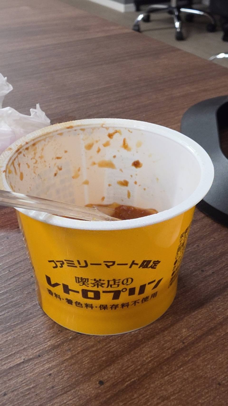 木目のデスクに置かれたファミリーマート限定の黄色いカップ入りレトロプリンです。中には食べかけのプリンとスプーンが見え、カップの内側にはカラメルの跡がついています。